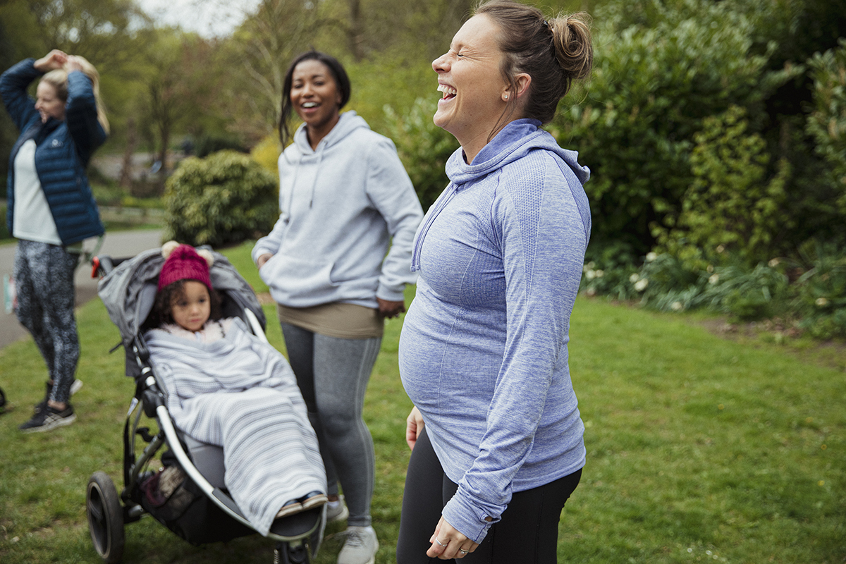 A group of women are standing in the park laughing and chatting together in the park with their pregnant fitness instructor.