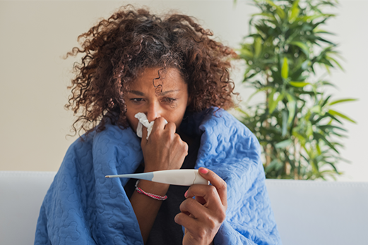 MVPDig0055_Flu Shots_Side Images_Stop the Spread_202508 a sick women wrapped in a blanket with a tissue looking at a thermometer