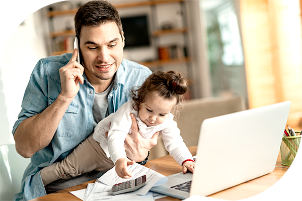 man holding infant while on mobile phone, sitting in front of a laptop.