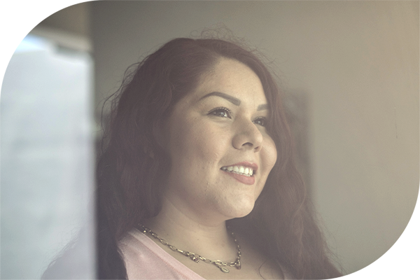 Smiling woman with long curly hair standing by a window, facing sideways and lit by natural daylight.