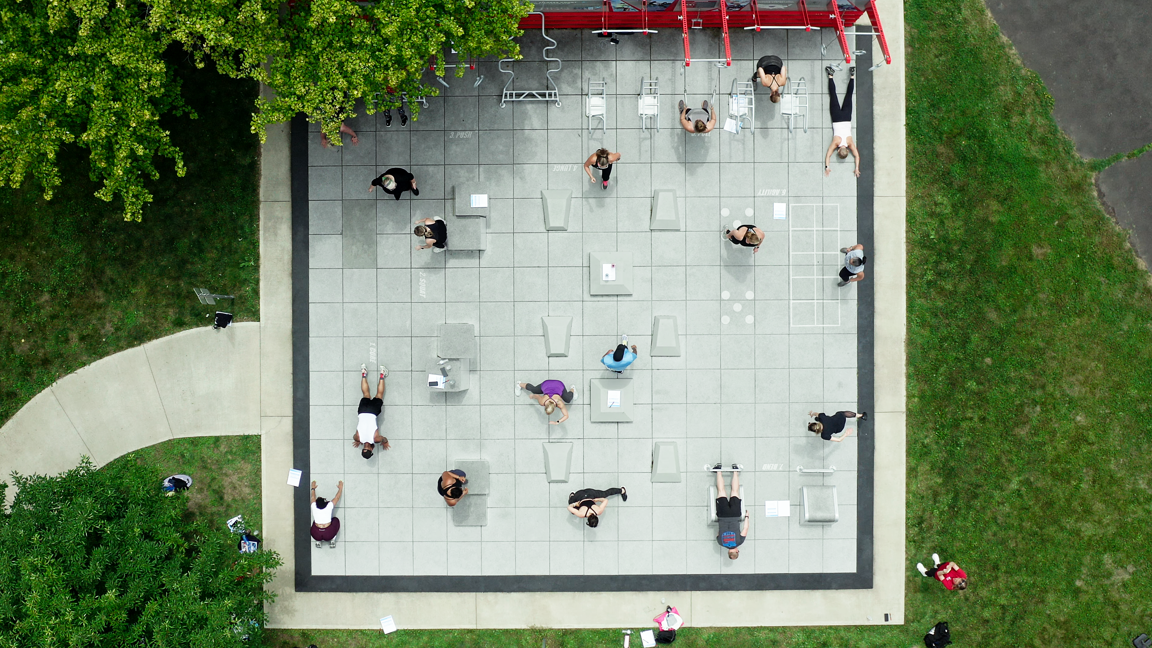 Aerial view of outdoor fitness area with people exercising at spaced stations on a tiled grid, surrounded by grass and trees.