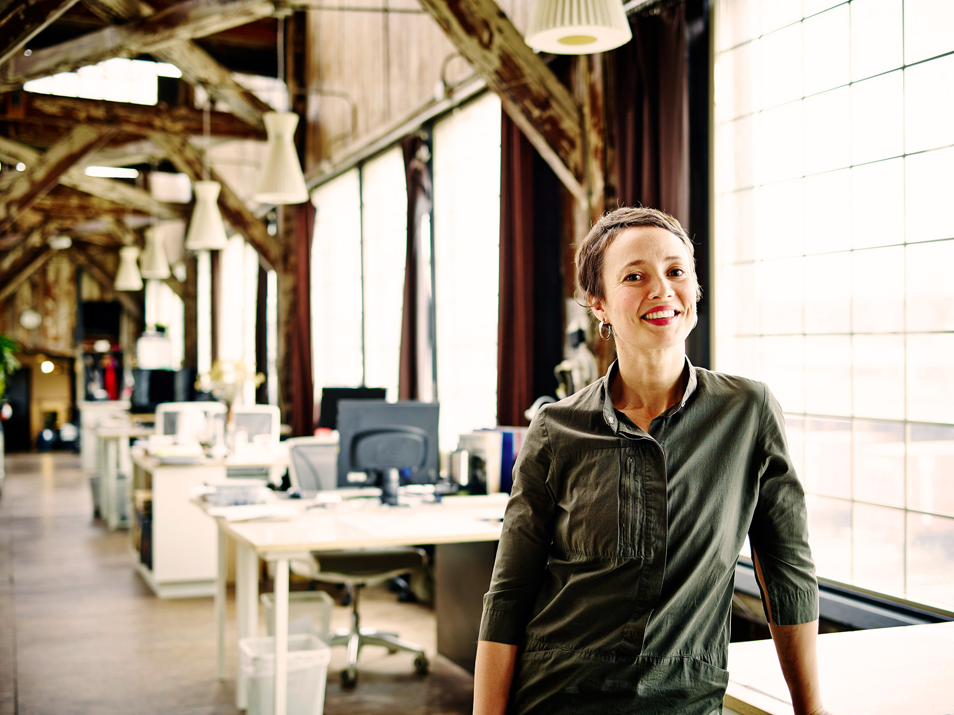A woman stands in an office beside a desk, looking engaged and ready to work