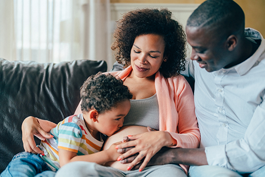 Child kissing a pregnant person's belly while sitting with family on a couch.
