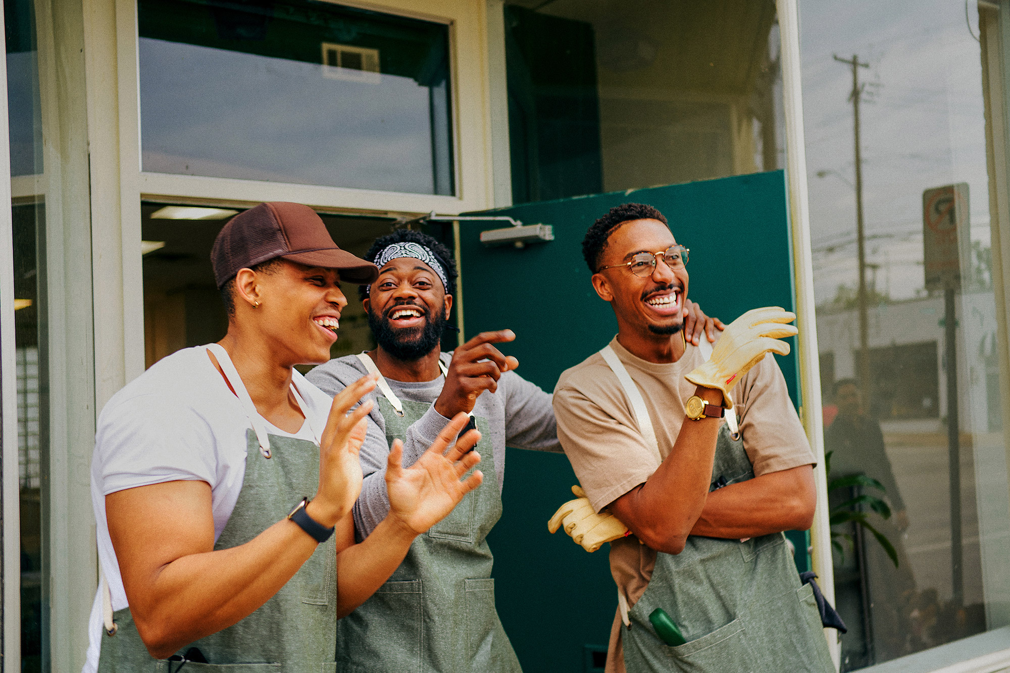 three-people-in-green-aprons-smiling-and-standing-outside-a-storefront Three people in green aprons smiling and standing outside a storefront.