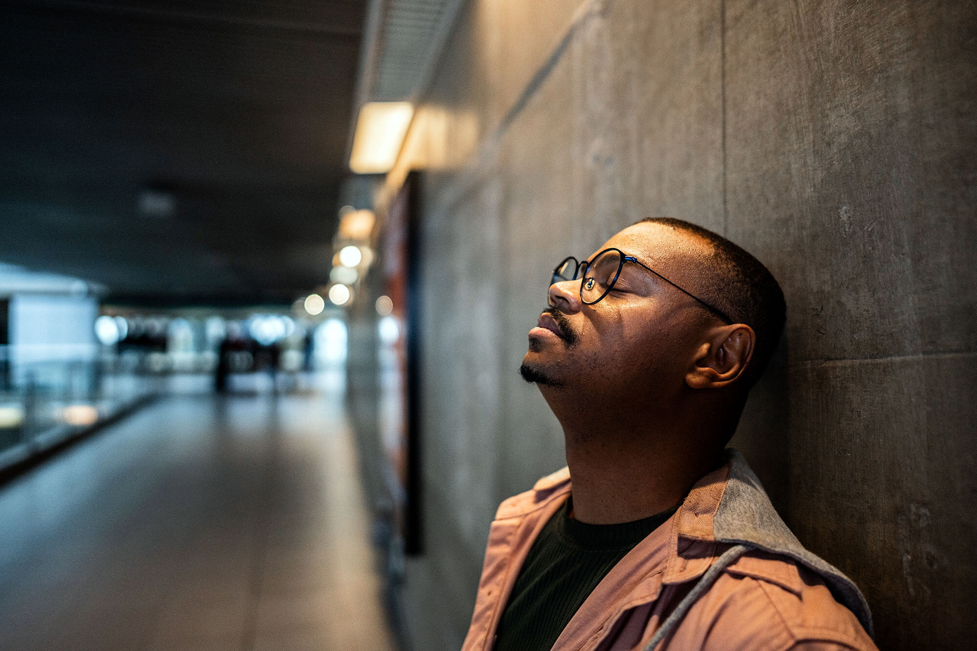 a man wearing glasses with his eye closed leans against a wall outdoors